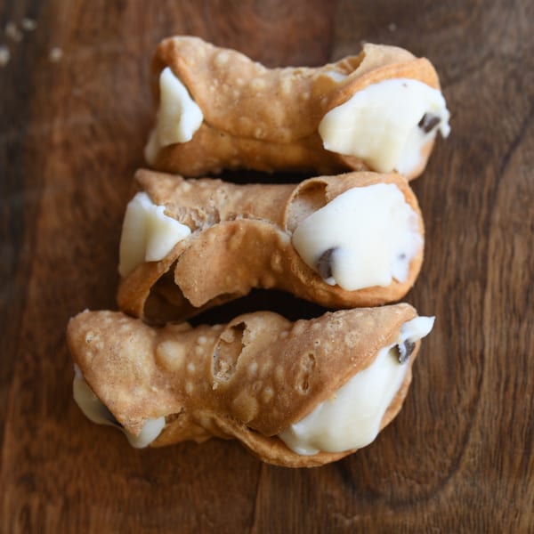 Three Louie's Pizza cannolis, filled with creamy white ricotta and chocolate chips, on a wooden surface.