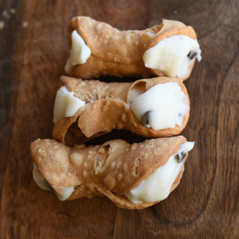 Three Louie's Pizza cannolis, filled with creamy white ricotta and chocolate chips, on a wooden surface.