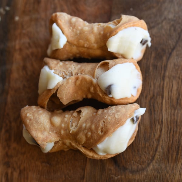 Three Louie's Pizza cannolis, filled with creamy white ricotta and chocolate chips, on a wooden surface.