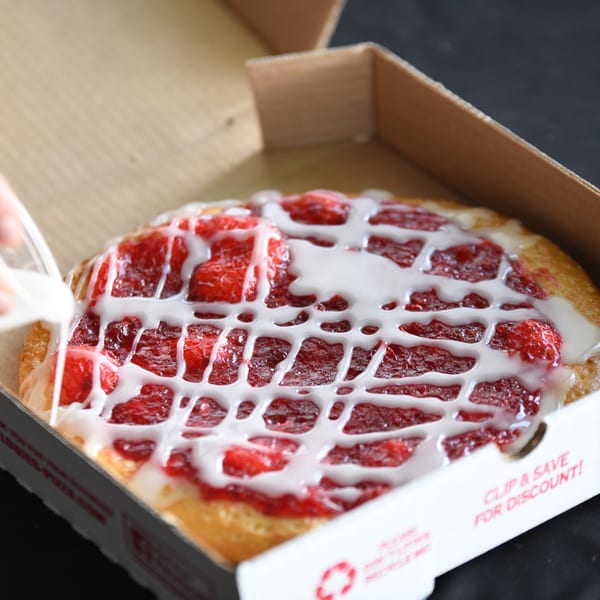 A hand drizzles white icing over a Louie's Pizza dessert pizza with strawberry topping in a box.