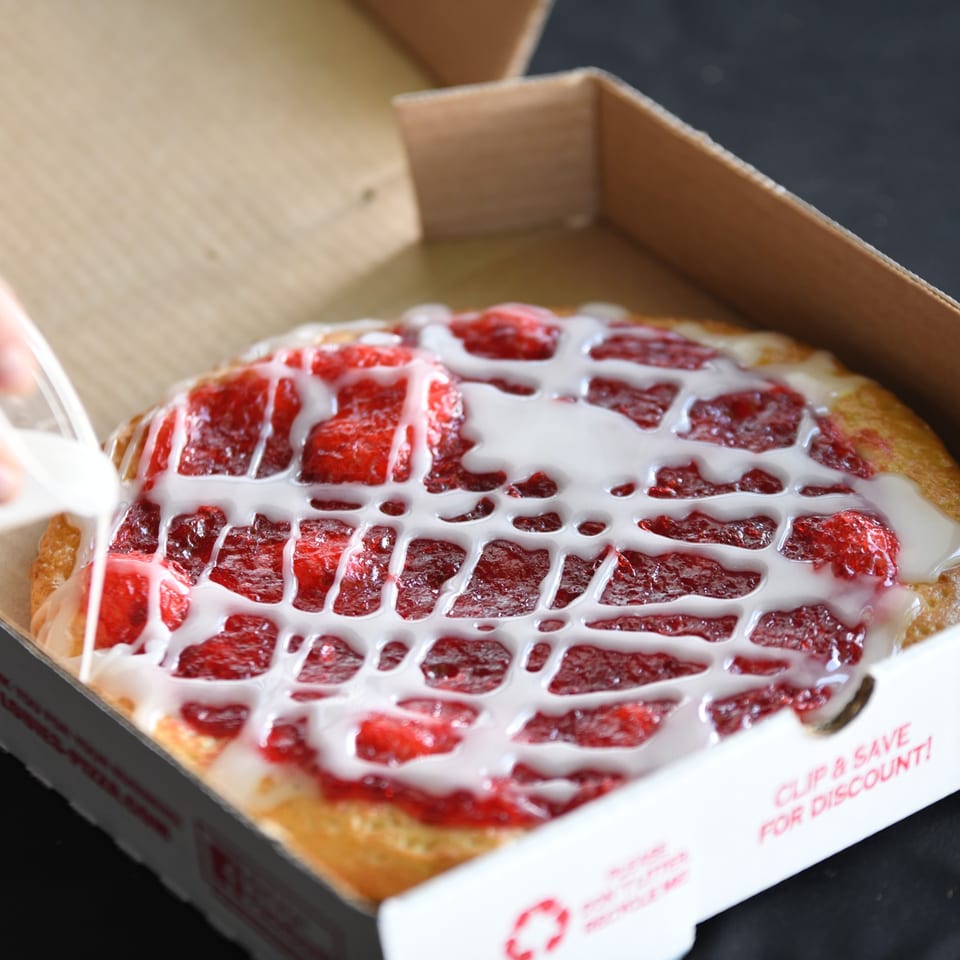 A hand drizzles white icing over a Louie's Pizza dessert pizza with strawberry topping in a box.