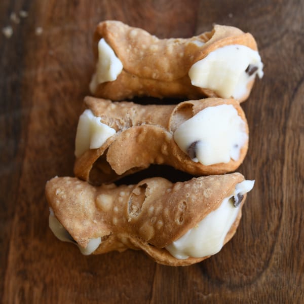 Three Louie's Pizza cannolis, filled with white cream and chocolate chips, on a wooden surface.