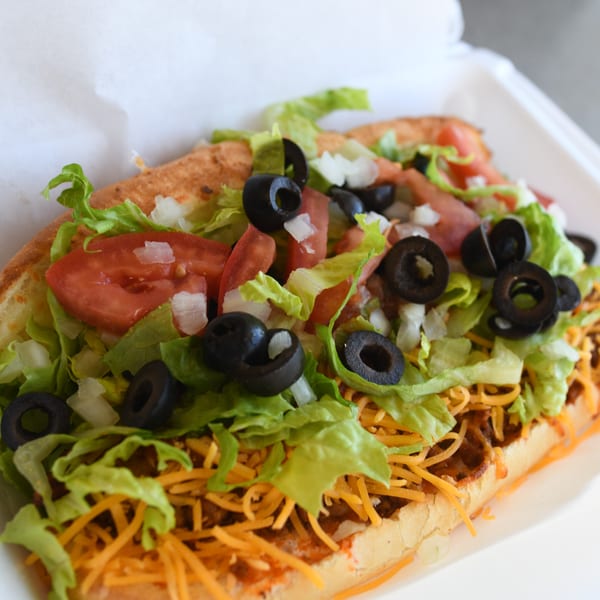 A Louie's Pizza taco sub with lettuce, tomatoes, black olives, and shredded cheese in a white container.