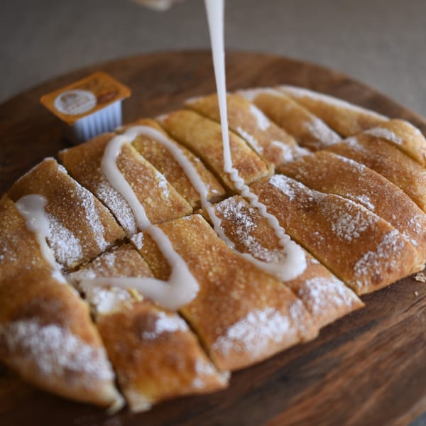 Sweet Louie's Pizza dessert breadsticks, drizzled with white icing and powdered sugar on a wooden board.